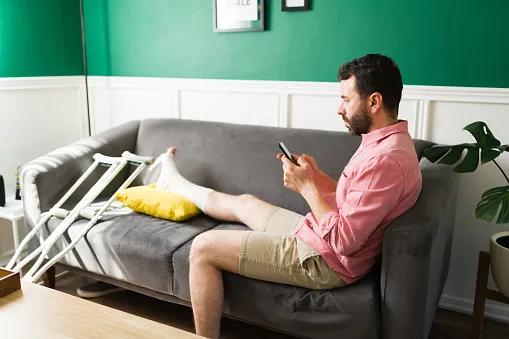 Man on the couch with crutches and a leg cast. He is looking at his phone and searching for a personal injury lawyer.